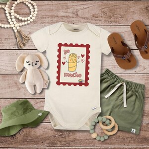 May include: Cream-colored baby onesie with a cartoon illustration inside a red postage stamp frame. The illustration features a smiling, yellow arepa with hearts and the text "yo mucho". Includes green shorts, sandals, a plush bunny, and a green hat.