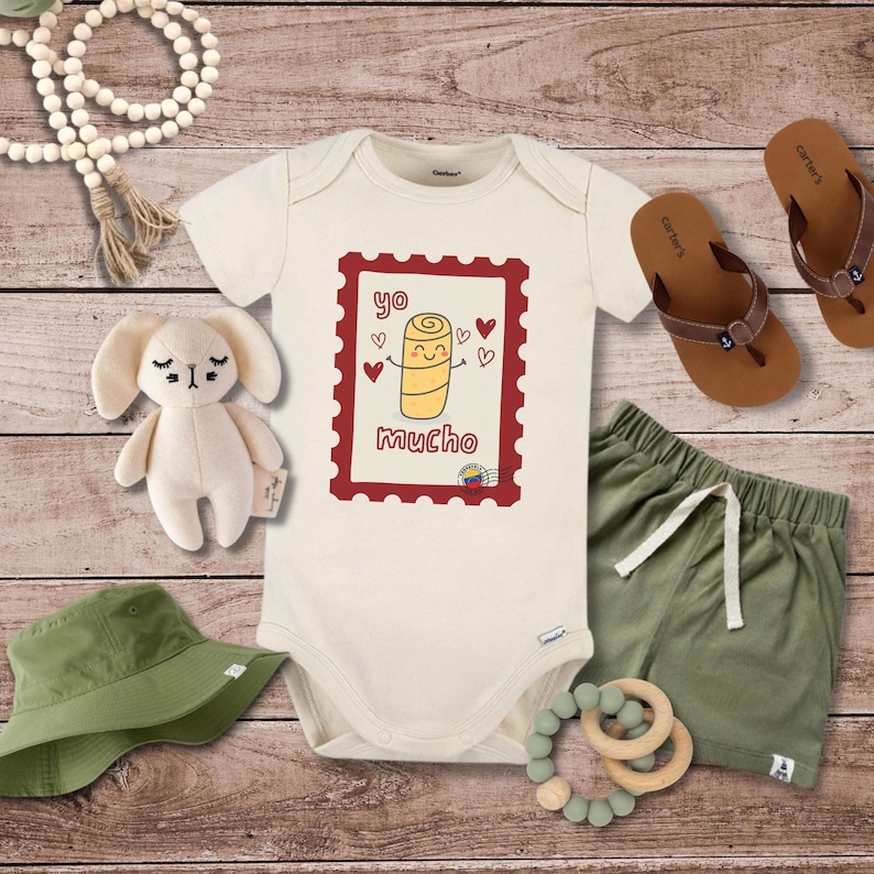 May include: Cream-colored baby onesie with a cartoon illustration inside a red postage stamp frame. The illustration features a smiling, yellow arepa with hearts and the text "yo mucho". Includes green shorts, sandals, a plush bunny, and a green hat.