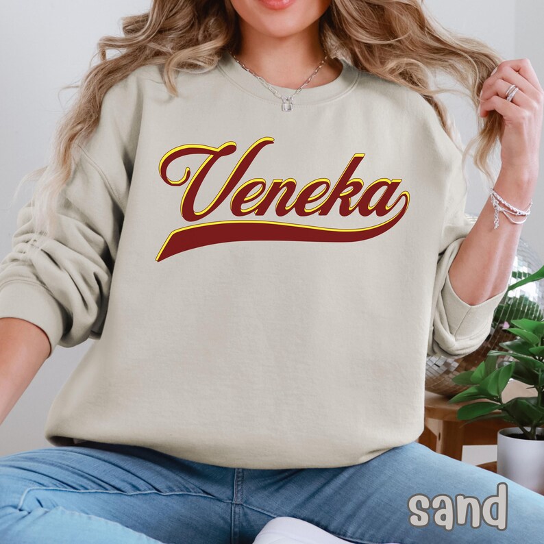 A person wearing a sand-colored crewneck sweatshirt with "Veneka" in red and yellow script across the chest, with "sand" written in white in the bottom right corner.