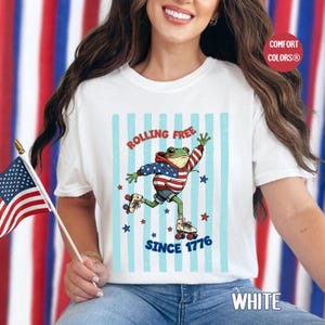 Pode incluir: Camiseta branca com um desenho de um sapo com um capuz patriótico e patins, com o texto "Rolling Free Since 1776". O design inclui listras azuis e estrelas. A camiseta tem o logotipo "Comfort Colors".