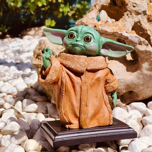 Puede incluir: Una figura verde de un personaje con orejas grandes y ojos grandes, que lleva una túnica marrón. La figura está sobre una base negra, rodeada de piedras blancas. El personaje está gesticulando con la mano.