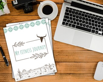 May include: A white journal with a turquoise and white patterned border. The journal has the text "My Fitness Journey" in a brown frame with a stylized bull skull graphic above the text. The journal is on a wooden surface with a laptop, a cup of coffee, a camera, and a mouse.