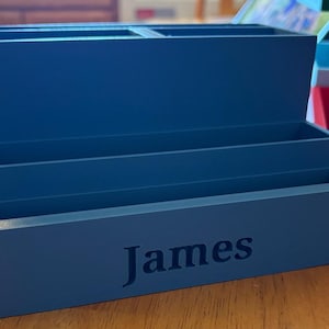 Könnte beinhalten: Ein blauer, abgestufter Schreibtisch-Organizer mit drei separaten Aufbewahrungsfächern in treppenartiger Anordnung. Auf der Vorderseite des untersten Fachs ist der Name James in dunkelblauer Schrift mittig aufgedruckt.