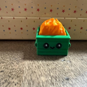 May include: A green 3D-printed miniature trash bin with a smiling face, holding an orange, crumpled object. The bin has small handles on the sides. The background is a wooden surface and a patterned box.