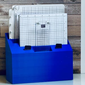 Peut inclure: Un organisateur en bois bleu vif contenant des outils d'estampage en acrylique transparent. Les outils comprennent une plateforme d'estampage quadrillée, une règle transparente et une plaque d'estampage blanche. L'organisateur a deux clips noirs et est posé sur un fond en bois.