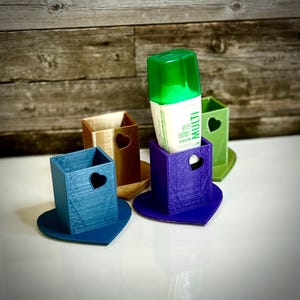 Op de afbeelding: Een verzameling van vier hartvormige penhouders in blauw, bruin, paars en groen. Elke houder heeft een hartvormige uitsparing en staat op een bijpassende hartvormige basis. Eén houder bevat een flesje vloeibare lijm.