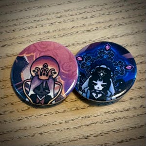 May include: Two circular badges with fantasy artwork. One has a pink and purple design of a crowned figure. The other features a character with a jeweled headdress against a blue backdrop, with pink gems.
