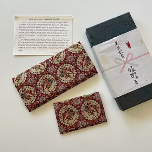 Könnte beinhalten: Eine rote und cremefarbene Stoffbörse mit einer passenden kleineren Börse, beide in einer schwarzen Schachtel mit einem weißen Etikett, auf dem in japanischen Schriftzeichen "未貝記念" steht. Die größere Börse hat eine Beschreibungskarte mit der Aufschrift "Lion Hunting Pattern Nishiki", die die Geschichte des Musters erklärt.