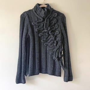 May include: A gray, long-sleeved sweater with a high collar and a zipper closure. The sweater features a unique, textured design on the front, with looped fabric creating an artistic pattern. The garment is hanging on a beige hanger.