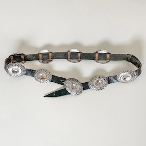 May include: A vintage black leather belt with silver-toned oval and square conchos. The belt features decorative metal accents and a classic buckle closure. The belt is a western-style accessory.