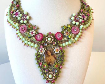 Puede incluir: Collar ornamentado con un colgante central ovalado que muestra un retrato vintage. El collar está adornado con flores de tela rosa, cuentas verdes y cristales. El diseño incorpora tonos verdes, rosas y dorados, creando una pieza llamativa.