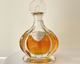 Può includere: Un flacone di profumo in vetro trasparente con un tappo decorativo. La bottiglia è riempita con un liquido color oro. L'etichetta recita "Eau de Possession Coro". La bottiglia è appoggiata su una superficie bianca.