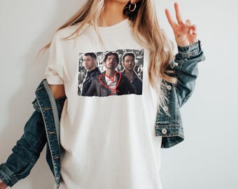 May include: Cream-colored t-shirt featuring a graphic of three men in jackets. The image is set against a backdrop of black and white images. The person in the middle has a red heart drawn on the image. The shirt is worn by a person wearing a denim jacket.