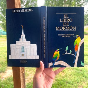 May include: A navy blue book with the title "EL LIBRO DE MORMON" and the text "OTRO TESTAMENTO DE JESUCRISTO". The cover features a painting of two colorful birds on a branch. The spine reads "EL LIBRO DE MORMON OTRO TESTAMENTO DE JESUCRISTO".