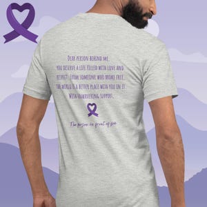 May include: A heather grey t-shirt with a purple ribbon and the text "DEAR PERSON BEHIND ME. YOU DESERVE A LIFE FILLED WITH LOVE AND RESPECT. FROM SOMEONE WHO BROKE FREE. THE WORLD IS A BETTER PLACE WITH YOU IN IT. WITH UNWAVERING SUPPORT. The person in front of you"