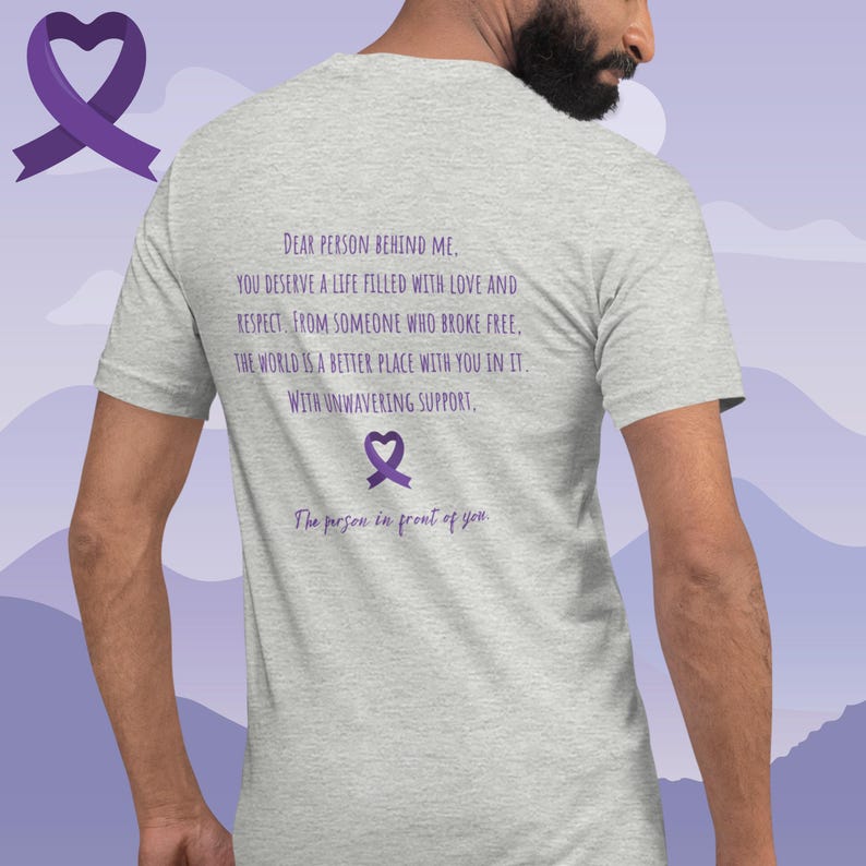 May include: A heather grey t-shirt with a purple ribbon and the text "DEAR PERSON BEHIND ME. YOU DESERVE A LIFE FILLED WITH LOVE AND RESPECT. FROM SOMEONE WHO BROKE FREE. THE WORLD IS A BETTER PLACE WITH YOU IN IT. WITH UNWAVERING SUPPORT. The person in front of you"