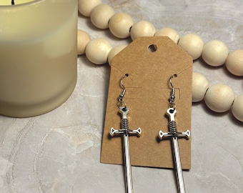 May include: A pair of silver sword earrings on a brown card. The swords have a detailed design and are hanging from silver hooks.