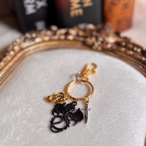 May include: A gold-coloured keyring featuring a black dragon charm, a small sword charm, and a decorative gold charm. The keyring is on a white surface with a gold-framed mirror in the background.