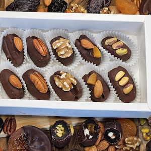 May include: A white box filled with individually wrapped chocolate-covered dates. Each date is topped with a different nut or dried fruit, including almonds, pecans, walnuts, pistachios, and dried apricots.