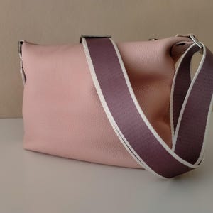 May include: A light pink leather crossbody bag with a wide, two-tone strap in purple and white. The bag has a rectangular shape and a silver-colored clasp. The strap is adjustable for shoulder or crossbody wear.