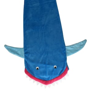 May include: A blue shark sleeping bag with a pink mouth and white teeth. The sleeping bag has a light blue tail fin and two light blue side fins.