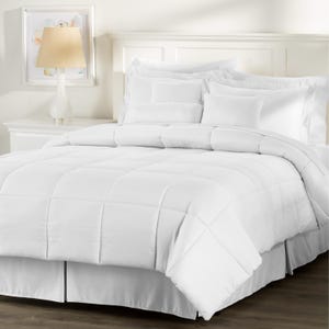 May include: A white bed set featuring a comforter, pillows, and a bed skirt. The comforter has a quilted pattern, and the pillows are various sizes. The bed is set against a white headboard and a white nightstand with a lamp.