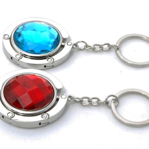 May include: Two purse hangers with a silver finish and a large, faceted gemstone. One has a bright blue gem, the other a deep red. Each has a chain and a keyring. These accessories are designed to hang a purse from a table.
