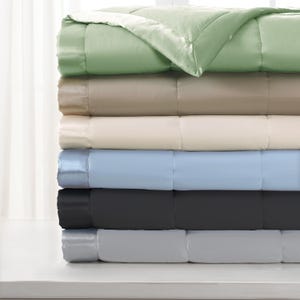 May include: A stack of six plush blankets in various colors, including green, beige, cream, light blue, dark gray, and light gray. Each blanket has a satin trim and a quilted design.