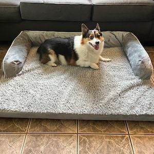 May include: A gray dog bed with a fluffy, gray, sherpa-like lining. The bed has a raised edge around the perimeter. A tri-colored dog is lying on the bed, looking directly at the camera.