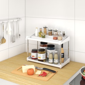 May include: White two-tiered kitchen shelf with jars, bottles, and spices. The shelf is on a countertop with a wooden cutting board, a tomato, and a knife.