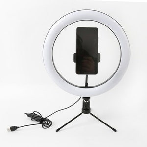 May include: A white ring light with a black smartphone holder and a black tripod stand. The ring light is illuminated, and a black smartphone is secured in the holder. A USB cable is connected to the base.