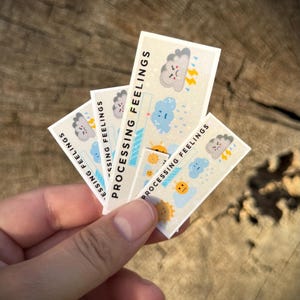 Może przedstawiać: Cztery prostokątne naklejki z napisem "PROCESSING FEELINGS" oraz ilustracjami chmur, deszczu i słońca. Naklejki trzymane są w dłoni na rozmytym drewnianym tle.