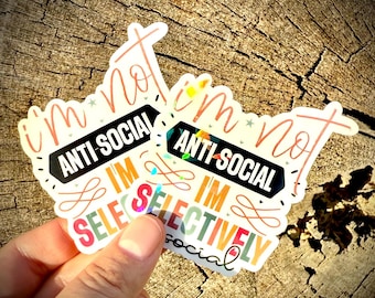 Op de afbeelding: Twee holografische stickers met de tekst "I'm not anti-social, I'm selectively social." De stickers zijn geplaatst tegen een houten achtergrond. De tekst is in verschillende kleuren, waaronder zwart, rood en oranje.