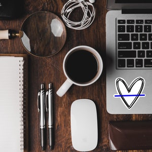 May include: A wooden desk with a laptop, a notebook, a magnifying glass, a cup of coffee, two pens, a wireless mouse, and a sticker of a heart with a blue line through it.
