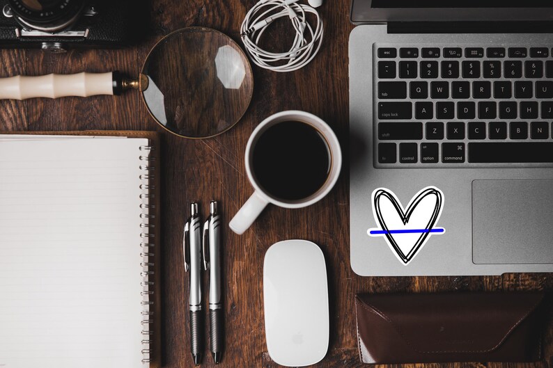 May include: A wooden desk with a laptop, a notebook, a magnifying glass, a cup of coffee, two pens, a wireless mouse, and a sticker of a heart with a blue line through it.