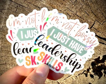 Op de afbeelding: Een kleurrijke sticker met de tekst "I'm not bossy, I just have leadership skills." De sticker heeft een geschulpte rand en heeft roze, teal, oranje en zwarte letters. Kleine hartjes en decoratieve elementen omringen de tekst.