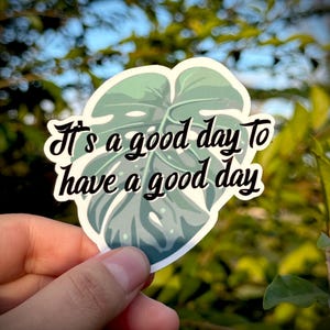 Puede incluir: Una pegatina con un diseño de hoja de monstera verde y el texto "It's a good day to have a good day" en escritura negra. La pegatina se sostiene en una mano sobre un fondo borroso de follaje verde y cielo azul.