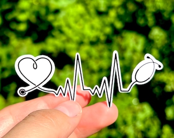 May include: A white sticker with a heart, a stethoscope, and a heartbeat line. The sticker is held in a hand against a green background.