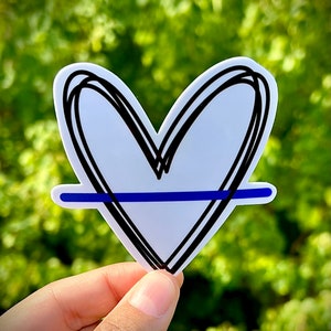 May include: A white heart sticker with a black outline and a thin blue line across the center. The sticker is held in a hand against a green background.
