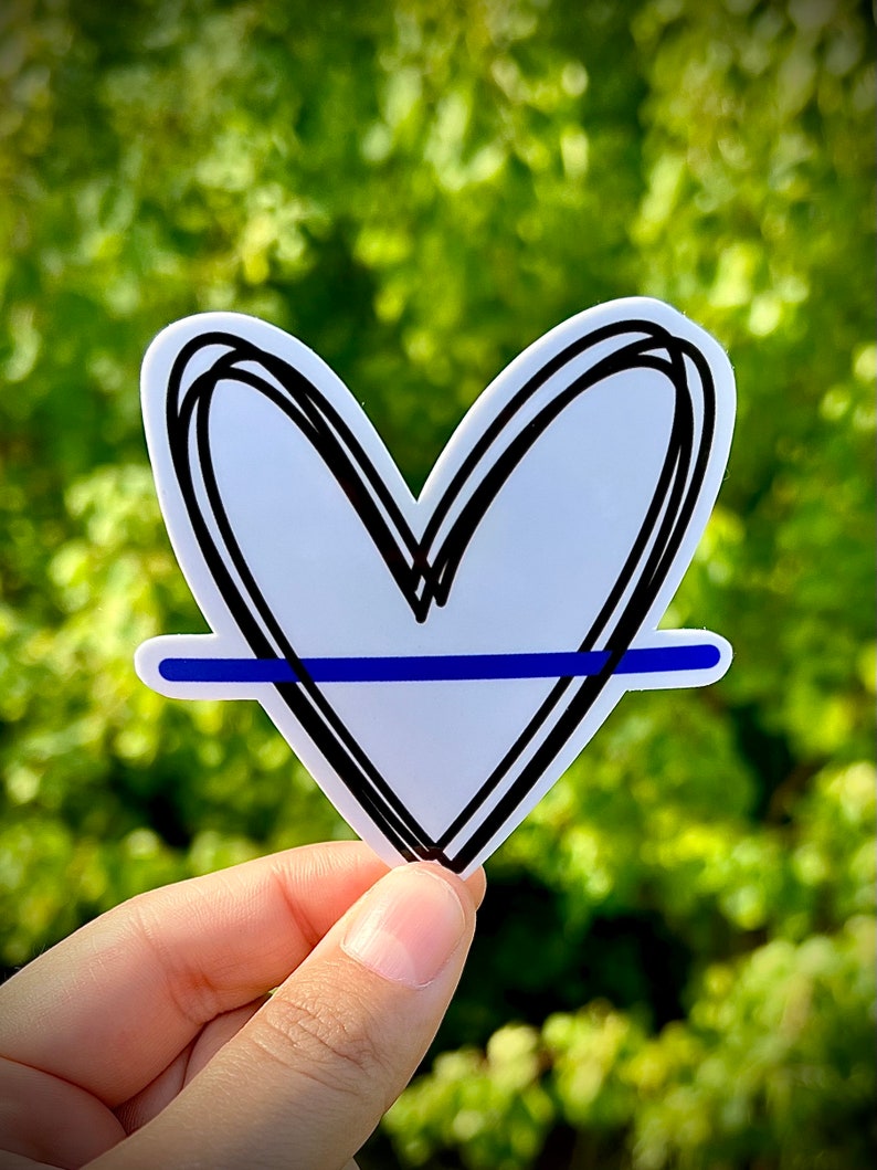 May include: A white heart sticker with a black outline and a thin blue line across the center. The sticker is held in a hand against a green background.