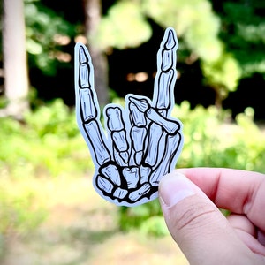 May include: A white sticker with a black outline of a skeleton hand making the rock and roll hand gesture.