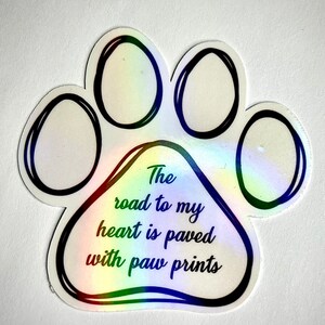 May include: A white paw print sticker with a rainbow iridescent finish. The sticker has black outlines and the text "The road to my heart is paved with paw prints".