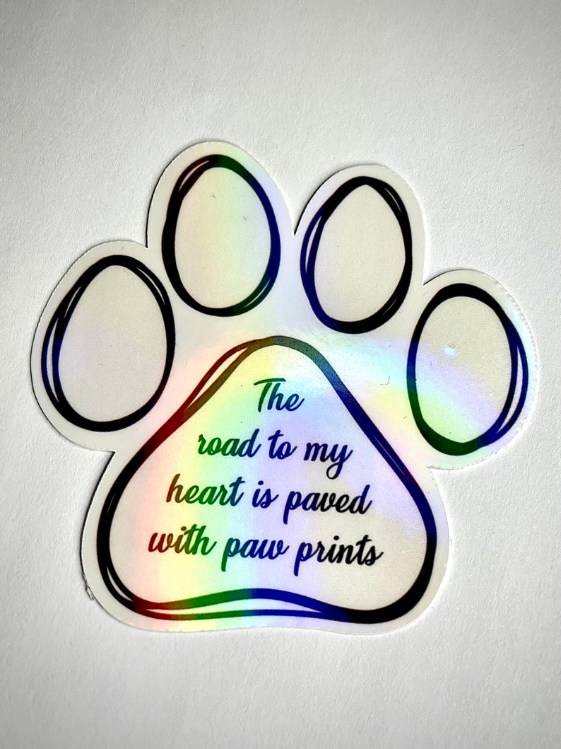 May include: A white paw print sticker with a rainbow iridescent finish. The sticker has black outlines and the text "The road to my heart is paved with paw prints".