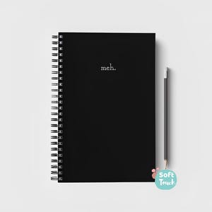 Può includere: Un quaderno nero a spirale con la parola "meh." stampata in bianco sulla copertina. Una matita nera è appoggiata accanto al quaderno.