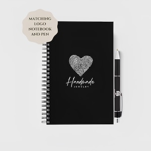 Op de afbeelding: Een zwart spiraalgebonden notitieboek en pennenset. Het notitieboek heeft een wit vingerafdruk hartontwerp en de woorden "Handmade Jewelry". De pen is zwart met zilveren details. Een sticker leest "Matching Logo Notebook and Pen".