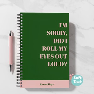 May include: A dark green spiral-bound notebook with a pink base and a pink pen. The notebook's cover displays the text "I'M SORRY, DID I ROLL MY EYES OUT LOUD?" in a light pink font. The notebook is on a white surface.