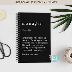 Op de afbeelding: Een zwart spiraalgebonden notitieboek met de definitie van "manager" in het wit gedrukt. Het notitieboek ligt op een witte ondergrond, met een kaars, schaar, een bol touw en groen.
