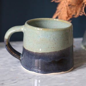 May include: A two-tone ceramic mug with a light blue-green upper section and a dark blue lower section. The mug features a curved handle and is set against a dark blue backdrop. The mug is likely handmade.