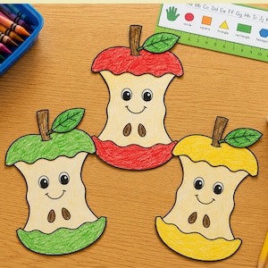 May include: Three paper cutouts of apple cores in red, green, and yellow, each featuring a hand-drawn smiling face, a brown stem, and a green leaf. The cutouts are arranged on a wooden surface alongside a blue crayon box, a ruler, and glue.