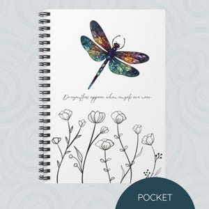 May include: A white spiral bound notebook with a colorful dragonfly illustration and the text "Dragonflies appear when angels are near". The notebook has a black and white line drawing of flowers on the bottom.
